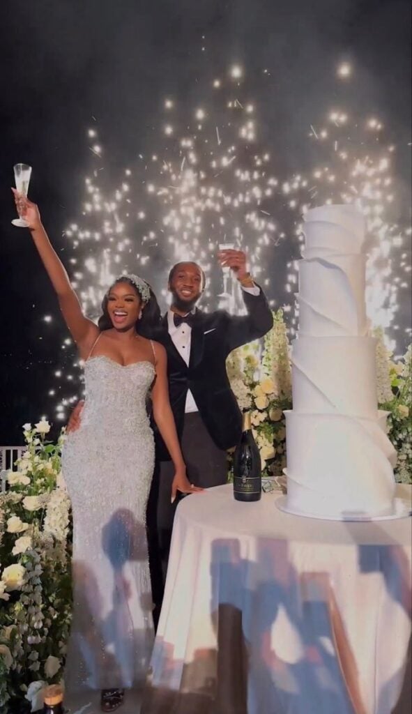 Joyful newlywed couple toasting with wine glasses while standing beside a tall wedding cake during their wedding celebration
