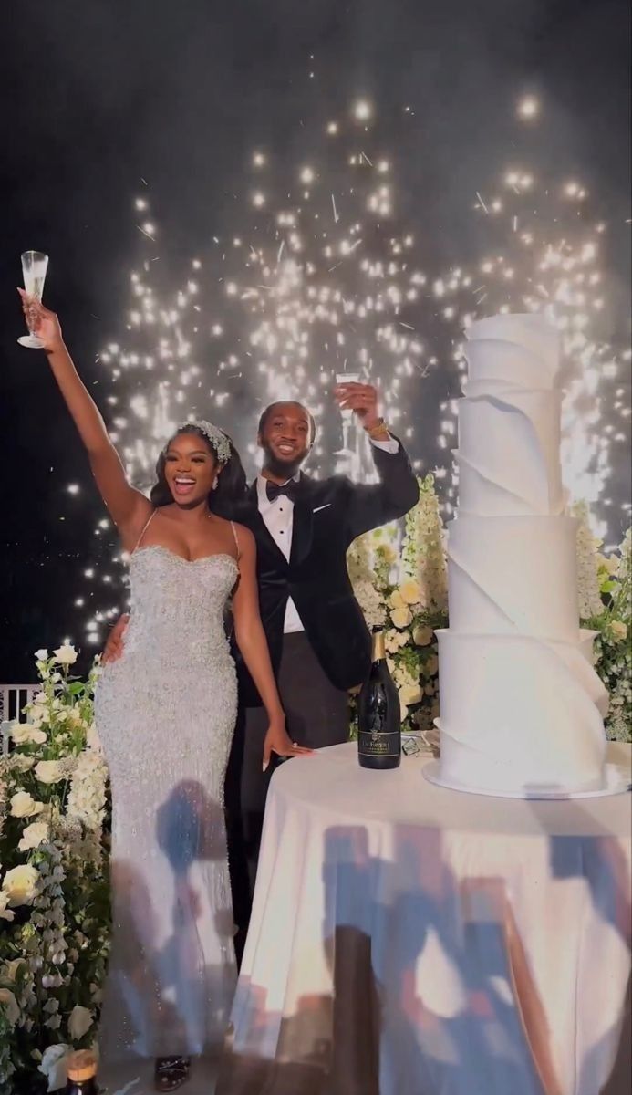 Joyful newlywed couple toasting with wine glasses while standing beside a tall wedding cake during their wedding celebration