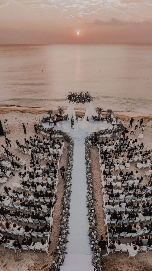 Elegant destination wedding ceremony on a beachfront venue at sunset with guests seated along a floral aisle
