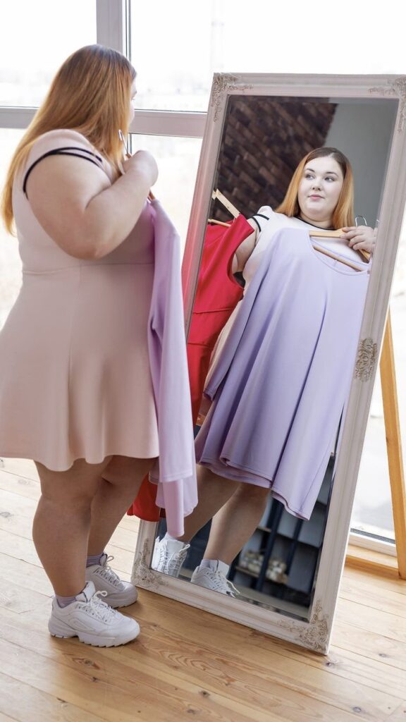 Plus-size woman standing in front of a mirror choosing an outfit before an event.