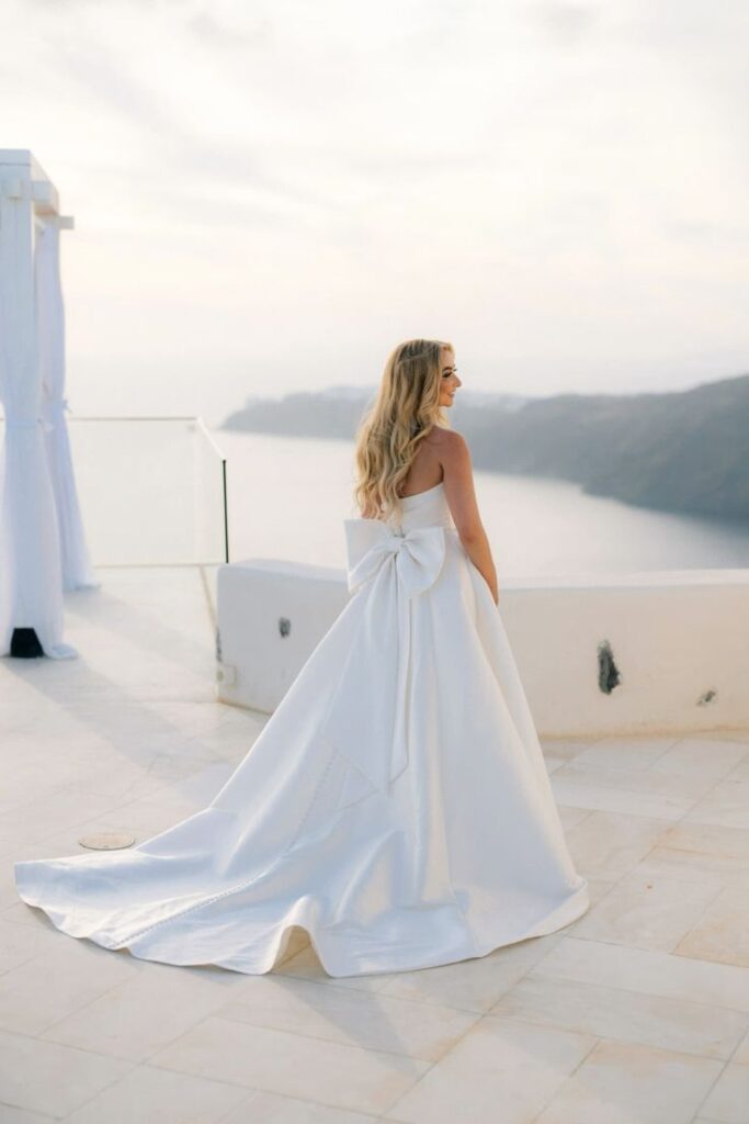 Bride wearing a satin A line wedding dress with a statement bow back and flowing train overlooking the sea.