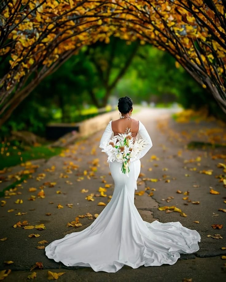 Bride in a white wedding dress walking beneath an autumn tree arch while holding a white floral bouquet.