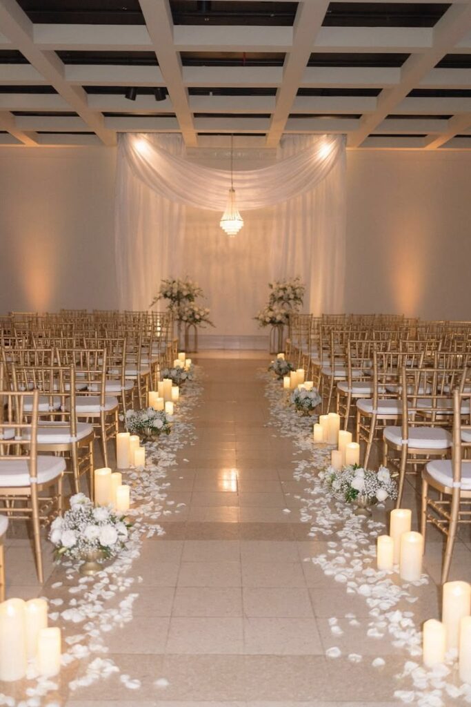 Elegant wedding ceremony aisle lined with glowing candles, white rose petals, and floral arrangements, leading to a draped altar with soft ambient lighting.
