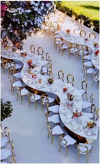 Elegant outdoor wedding reception featuring curved banquet tables with white linens, gold chairs, floral centerpieces, and a lush hanging flower installation for a luxurious garden celebration.