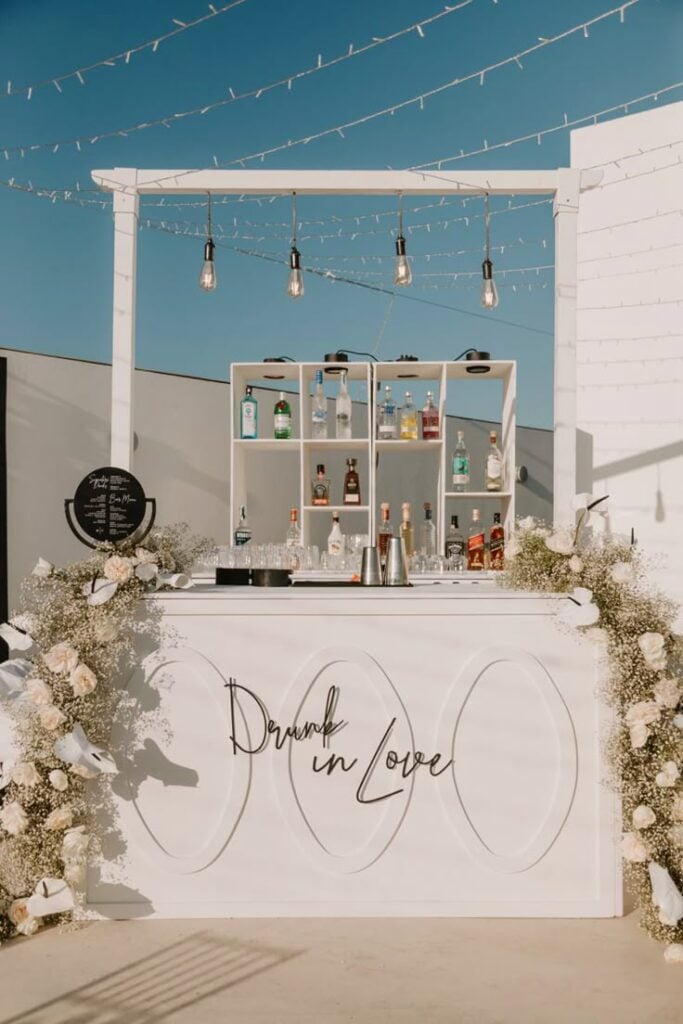 Elegant outdoor wedding bar with a white custom counter, black script signage, floral arrangements in white blooms and baby's breath, open shelving with drinks, and string lights overhead.