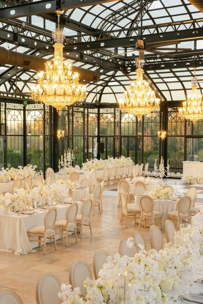Elegant European-inspired wedding reception in a glass conservatory featuring crystal chandeliers, white floral centerpieces, candlelit tables, classic French-style chairs, and timeless luxury décor.