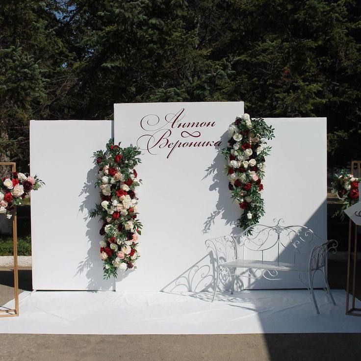 Elegant wedding photo backdrop with a white display wall, cascading red and white floral arrangements, personalized calligraphy, and a decorative white bench for guest photos and portraits.