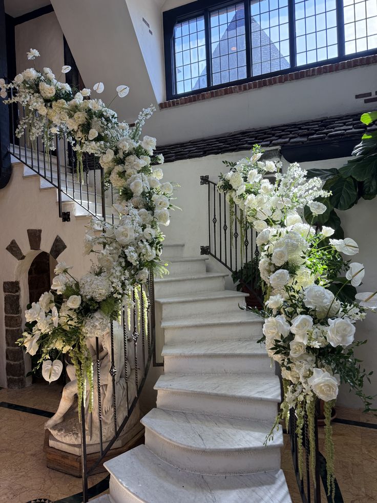 Elegant staircase decorated with lush white wedding flowers, roses, hydrangeas, anthuriums, cascading greenery, and floral garlands wrapped around the railings for a luxurious indoor wedding entrance.