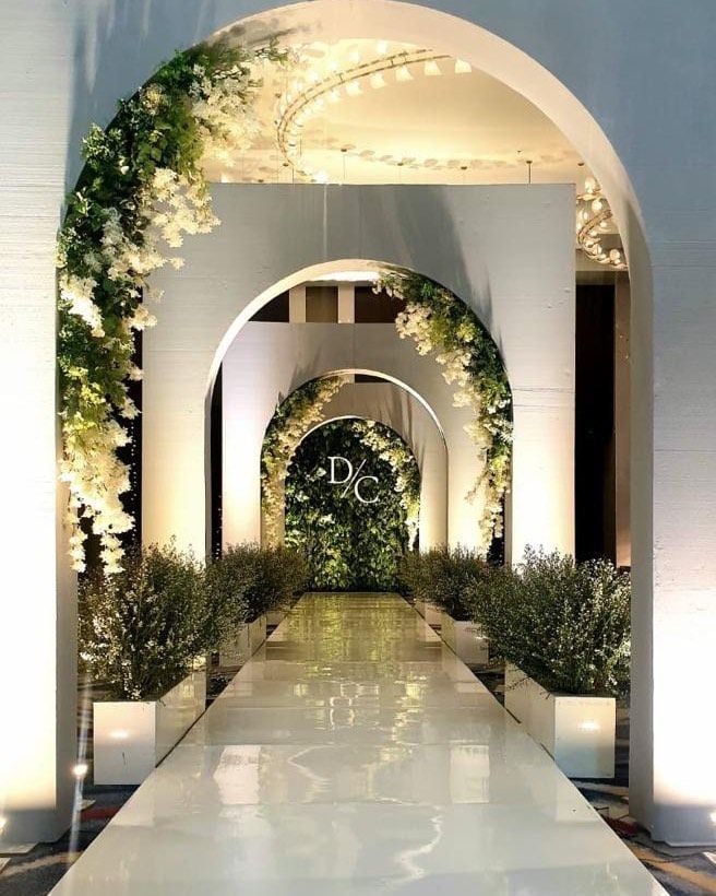 Luxury wedding entrance featuring elegant white arches adorned with cascading white flowers and greenery, a glossy aisle, soft ambient lighting, and lush plants creating a grand ceremony walkway.