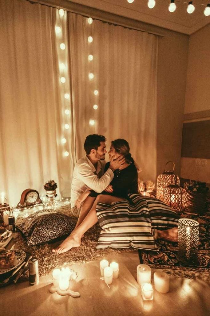 Couple sharing a romantic candlelit evening indoors, surrounded by fairy lights, cozy cushions, and warm ambient dรฉcor.