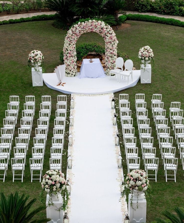 Elegant romantic garden wedding ceremony setup with a white aisle, blush and white floral arch, matching flower arrangements, white ceremony chairs, and lush greenery creating a timeless outdoor wedding theme.