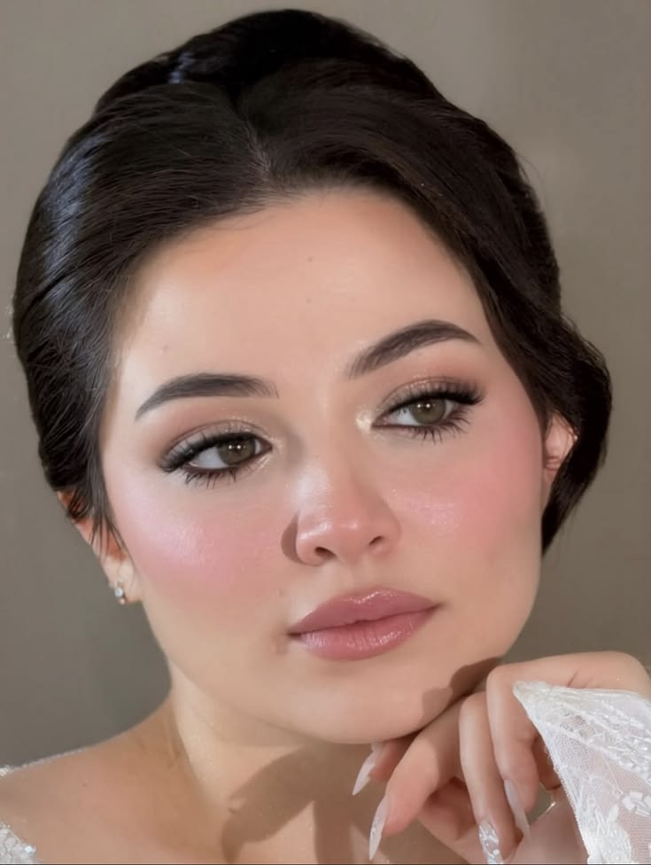 Bride with satin-finish makeup featuring soft taupe eyeshadow, rosy blush, glowing skin, fluttery lashes, glossy nude lips, and an elegant swept-back updo.