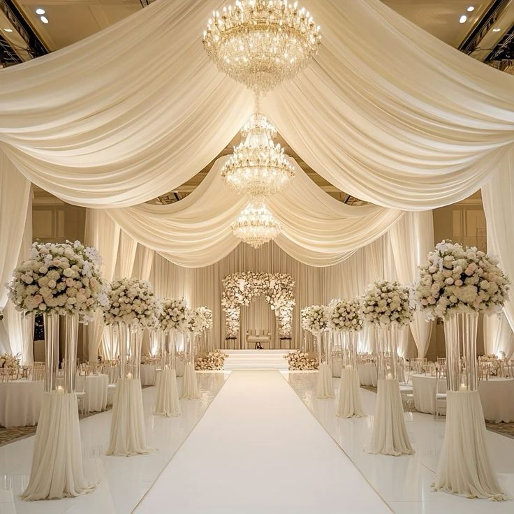 Luxurious wedding venue with flowing white ceiling drapes, crystal chandeliers, tall white floral arrangements, and a grand aisle leading to an elegant floral ceremony backdrop.