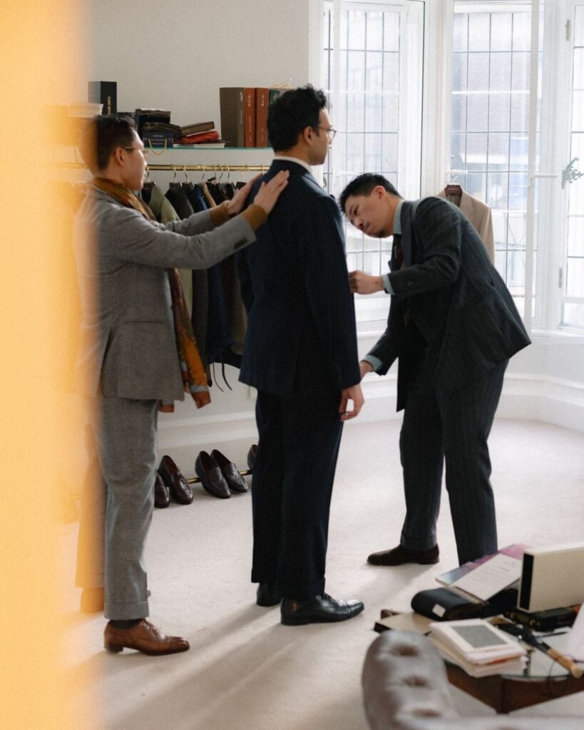 Groom trying on a tailored wedding suit while stylists make final fitting adjustments in a tailoring studio.