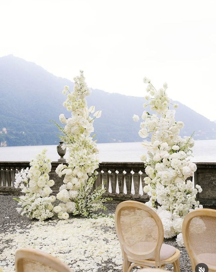 Elegant outdoor wedding ceremony setup with tall white floral arrangements, petal-lined aisle, and scenic lakeside mountain views.