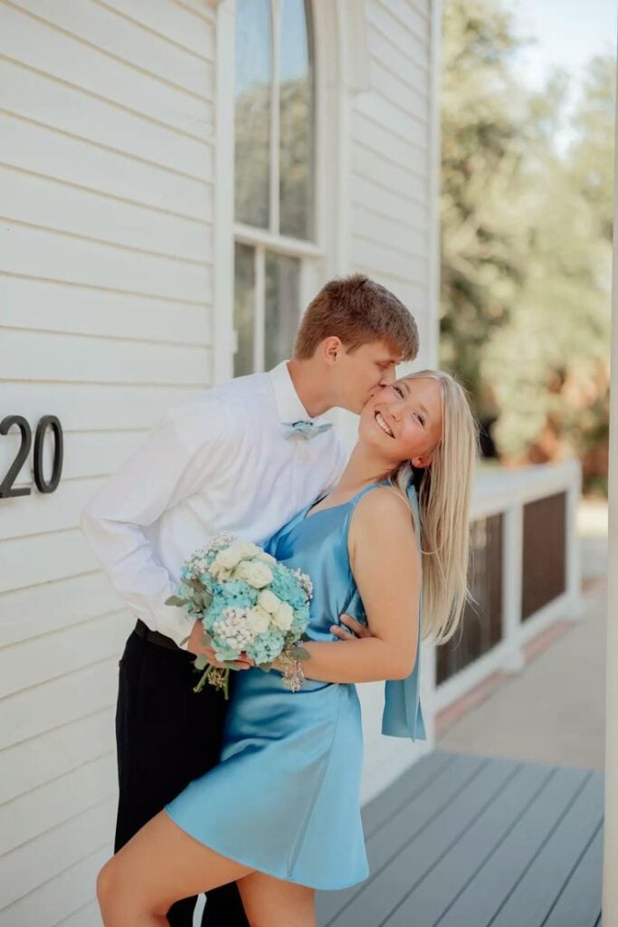 Happy engaged couple embracing outdoors, with the groom kissing his fiancรฉe on the cheek as she holds a bouquet of blue and white flowers.
