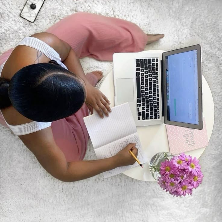 A lady sitting on the floor with her laptop on a small table placed in front of her, she is writing on a notebook