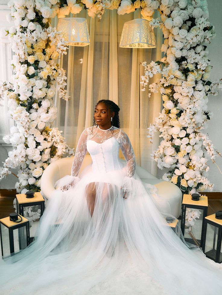 Bride wearing a long sleeve lace wedding dress with a flowing tulle skirt, seated beneath an elegant white floral arch.